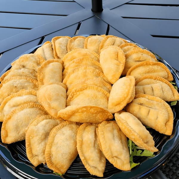 A large platter of golden-brown Catania's Pizza empanadas, garnished with fresh greens, on a dark table.