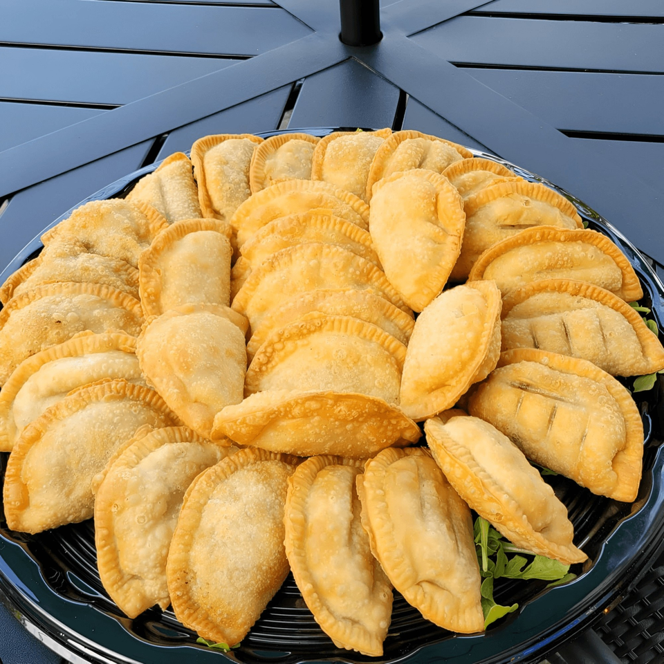 A large platter of golden-brown Catania's Pizza empanadas, garnished with fresh greens, on a dark table.