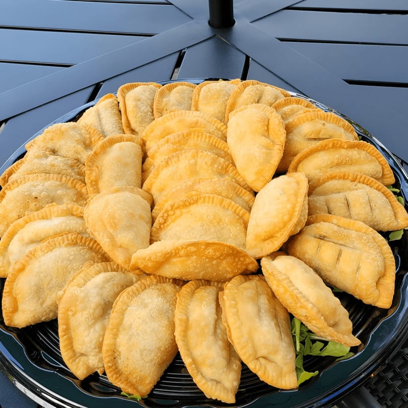 A large platter of golden-brown Catania's Pizza empanadas, garnished with fresh greens, on a dark table.