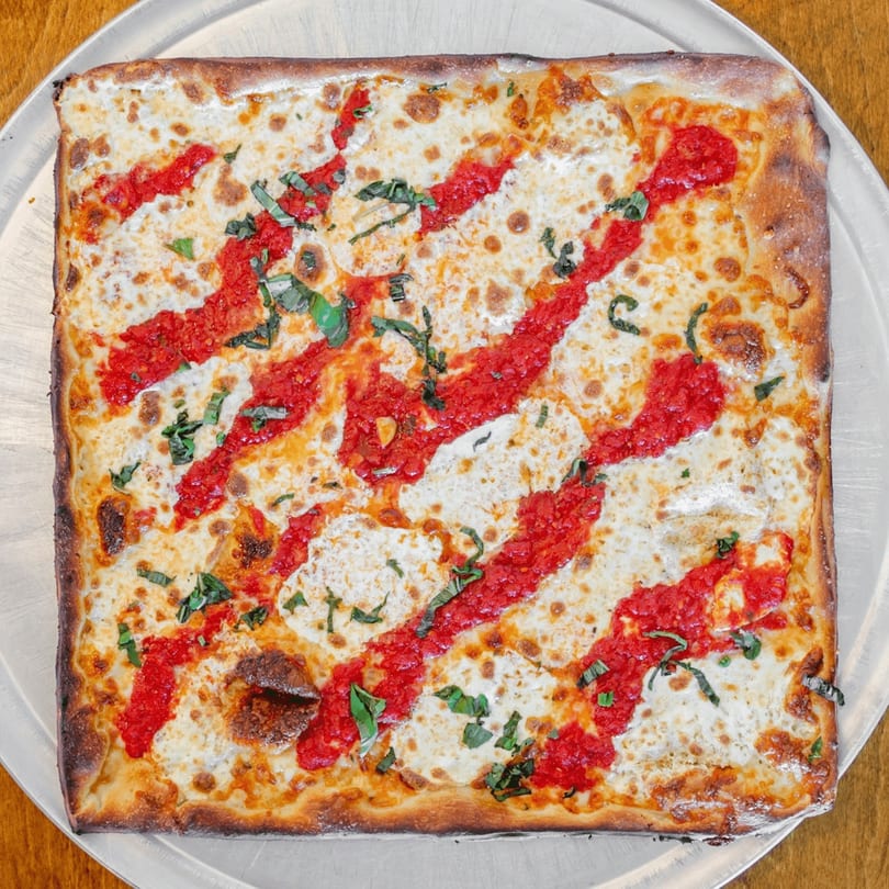 Square Catania's Pizza with red sauce stripes, melted cheese, and fresh basil on a metal tray.